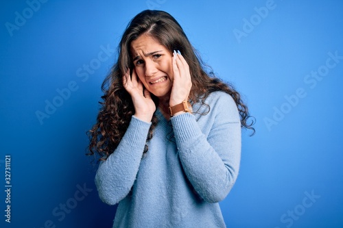 Young beautiful woman with curly hair wearing blue casual sweater over isolated background covering ears with fingers with annoyed expression for the noise of loud music. Deaf concept.