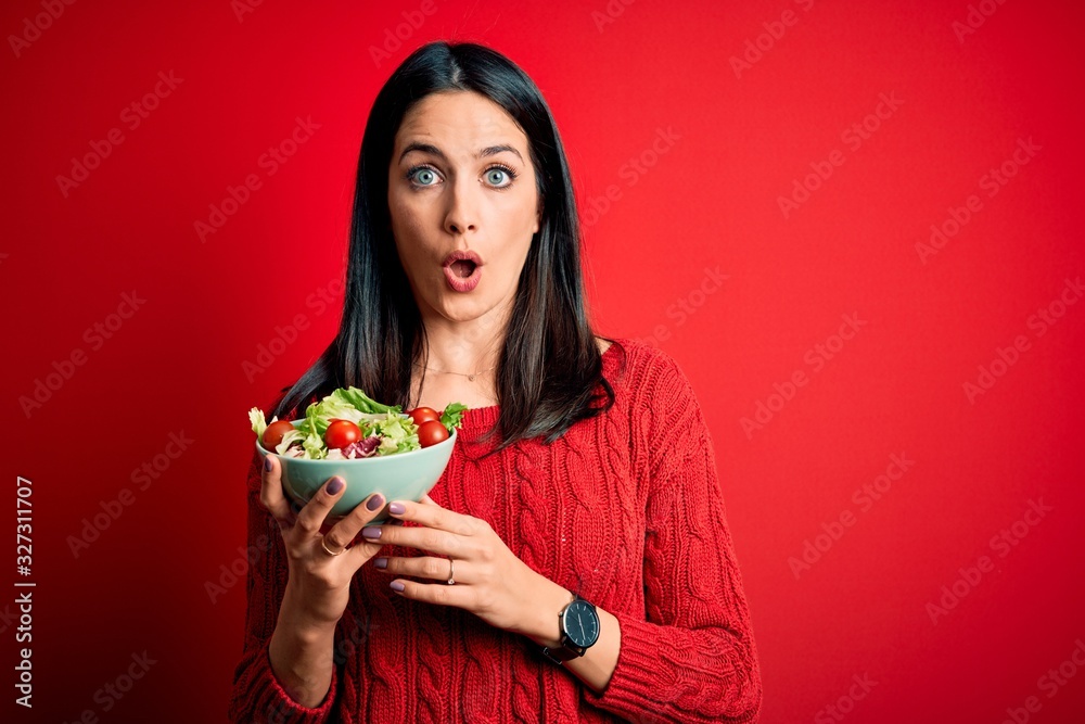 Young brunette woman with blue eyes eating healthy green salad over isolated red background scared in shock with a surprise face, afraid and excited with fear expression Young brunette woman with blue eyes eating healthy green salad over isolated red background scared in shock with a surprise face, afraid and excited with fear expression