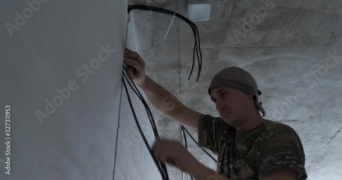 At construction site, male electrician attaches electric cables to concrete ceiling, repairs in new building, electrical network, wiring, man in building uniform, cable man, qualified electrician