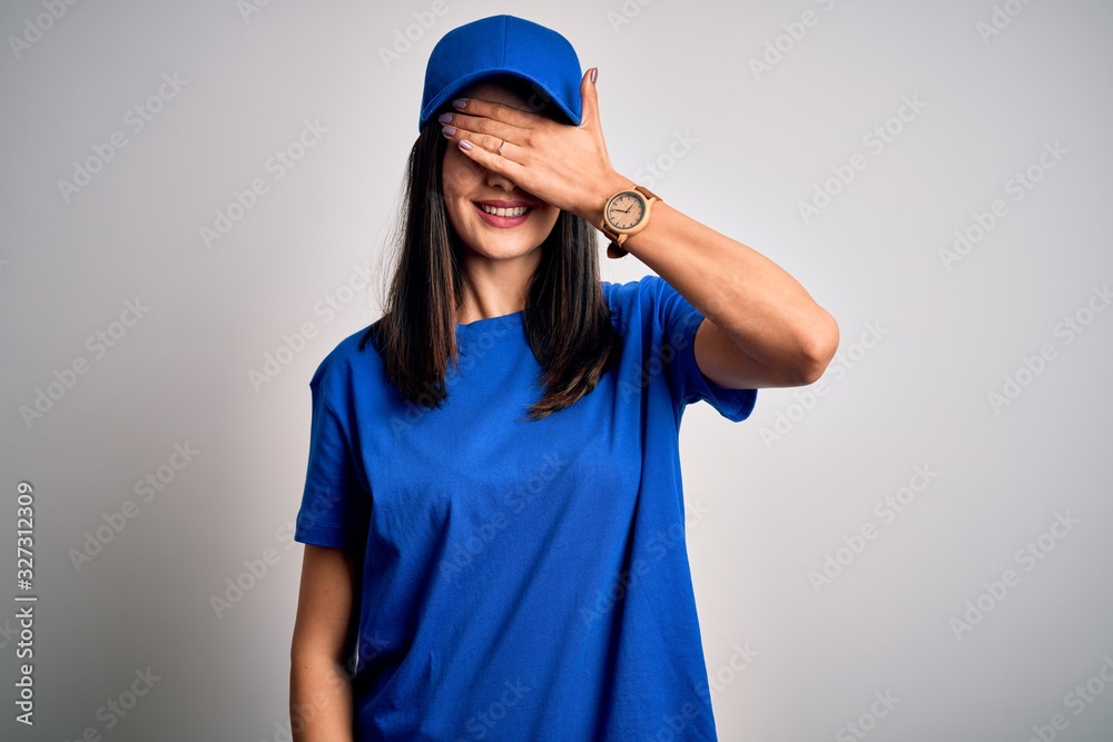 Young delivery woman with blue eyes wearing cap standing over blue background smiling and laughing with hand on face covering eyes for surprise. Blind concept. Young delivery woman with blue eyes wearing cap standing over blue background smiling and laughing with hand on face covering eyes for surprise. Blind concept.