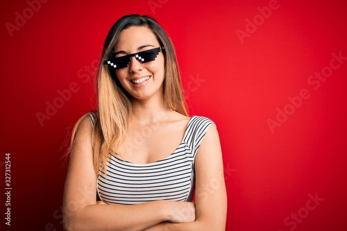 Young beautiful brunette woman wearing funny thug life sunglasses over red background happy face smiling with crossed arms looking at the camera. Positive person.