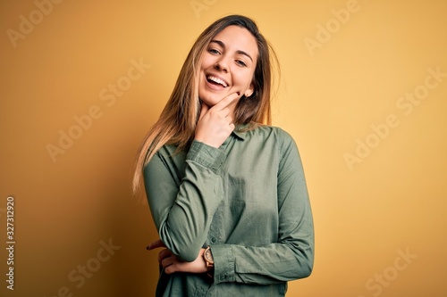 Young beautiful blonde woman with blue eyes wearing green shirt over yellow background looking confident at the camera smiling with crossed arms and hand raised on chin. Thinking positive. © Krakenimages.com