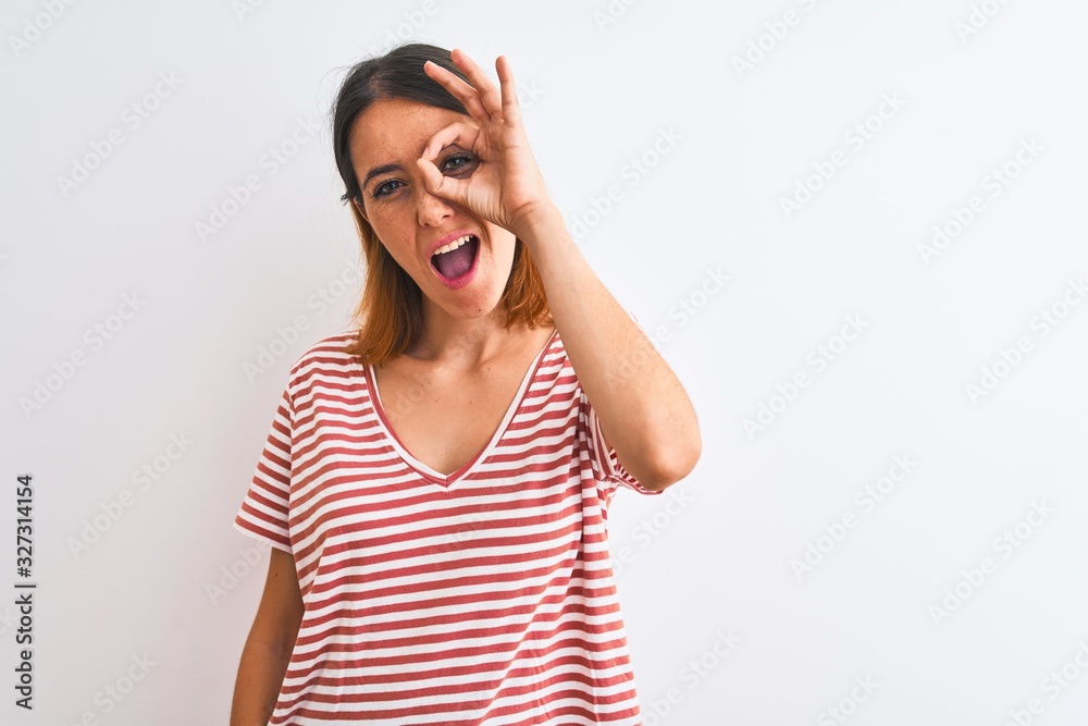 Beautiful redhead woman wearing casual striped red t-shirt over isolated background doing ok gesture with hand smiling, eye looking through fingers with happy face. Beautiful redhead woman wearing casual striped red t-shirt over isolated background doing ok gesture with hand smiling, eye looking through fingers with happy face.