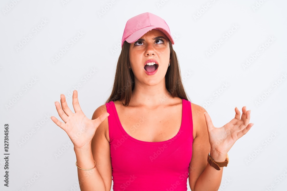 Young beautiful girl wearing pink casual t-shirt and cap over isolated white background crazy and mad shouting and yelling with aggressive expression and arms raised. Frustration concept. Young beautiful girl wearing pink casual t-shirt and cap over isolated white background crazy and mad shouting and yelling with aggressive expression and arms raised. Frustration concept.