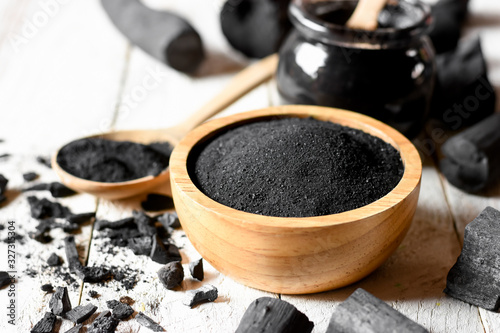 Black charcoal powder for facial mask and scrub, placed on a white wooden table, health and beauty concept.