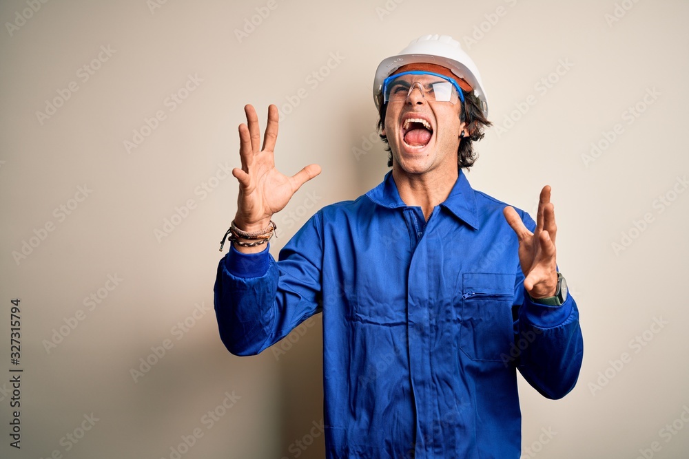 Young constructor man wearing uniform and security helmet over isolated white background crazy and mad shouting and yelling with aggressive expression and arms raised. Frustration concept.