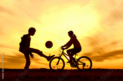 Girl riding bike in countryside  boy playing with ball outdoors  silhouette of riding and playing  persons at sunset in nature