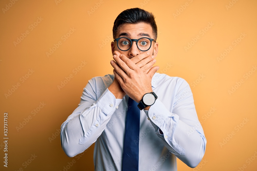 Young handsome businessman wearing tie and glasses standing over yellow background shocked covering mouth with hands for mistake. Secret concept. Young handsome businessman wearing tie and glasses standing over yellow background shocked covering mouth with hands for mistake. Secret concept.