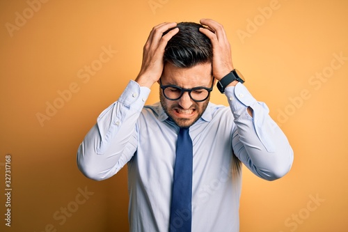 Young handsome businessman wearing tie and glasses standing over yellow background suffering from headache desperate and stressed because pain and migraine. Hands on head.