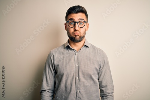 Young handsome man wearing elegant shirt and glasses over isolated white background puffing cheeks with funny face. Mouth inflated with air, crazy expression. © Krakenimages.com