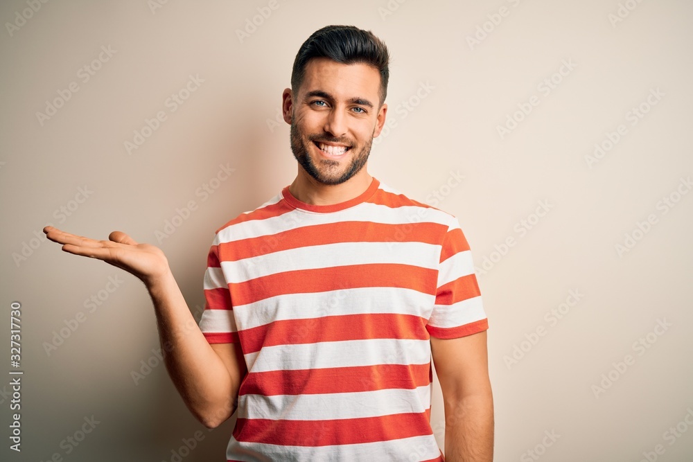 Young handsome man wearing casual striped t-shirt standing over isolated white background smiling cheerful presenting and pointing with palm of hand looking at the camera. Young handsome man wearing casual striped t-shirt standing over isolated white background smiling cheerful presenting and pointing with palm of hand looking at the camera.