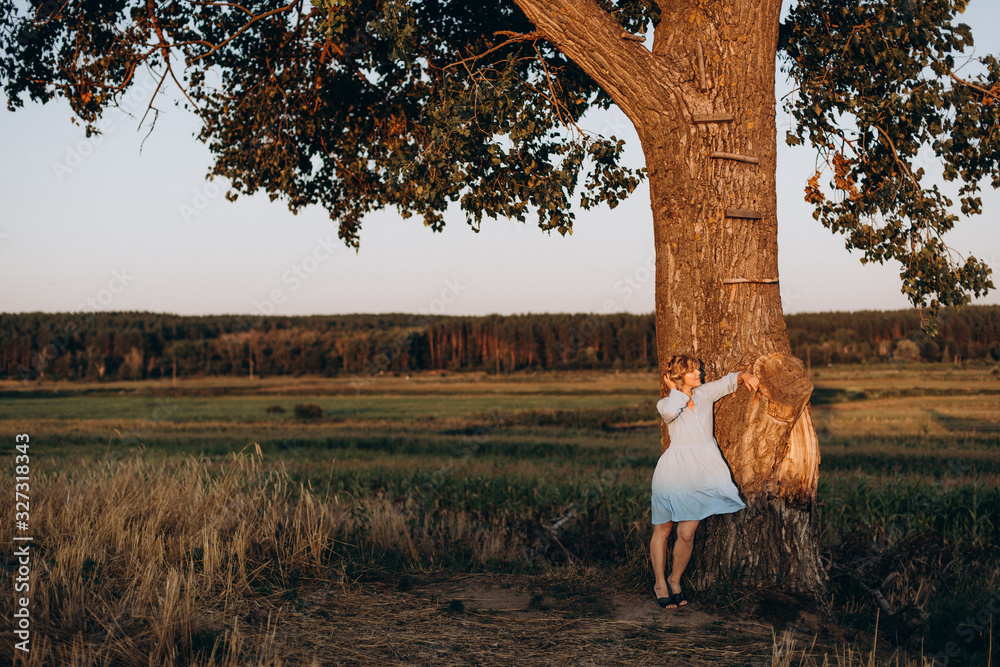 Beautiful free woman in a light summer dress in a field near a large tree at sunset. Soft selective focus.