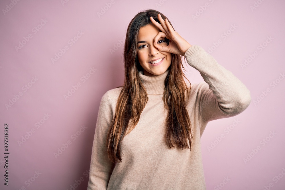Young beautiful girl wearing casual turtleneck sweater standing over isolated pink background doing ok gesture with hand smiling, eye looking through fingers with happy face.