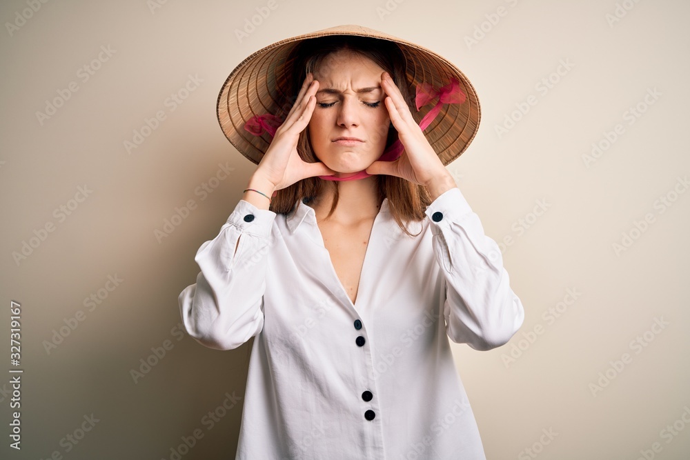 Young beautiful redhead woman wearing asian traditional conical hat over white background suffering from headache desperate and stressed because pain and migraine. Hands on head. Young beautiful redhead woman wearing asian traditional conical hat over white background suffering from headache desperate and stressed because pain and migraine. Hands on head.