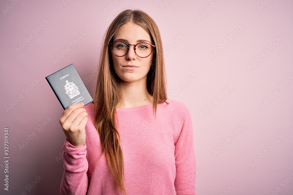 Beautiful redhead tourist woman holding canadian canada passport over pink bakcground with a confident expression on smart face thinking serious