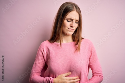 Young beautiful redhead woman wearing casual sweater over isolated pink background with hand on stomach because indigestion, painful illness feeling unwell. Ache concept. © Krakenimages.com