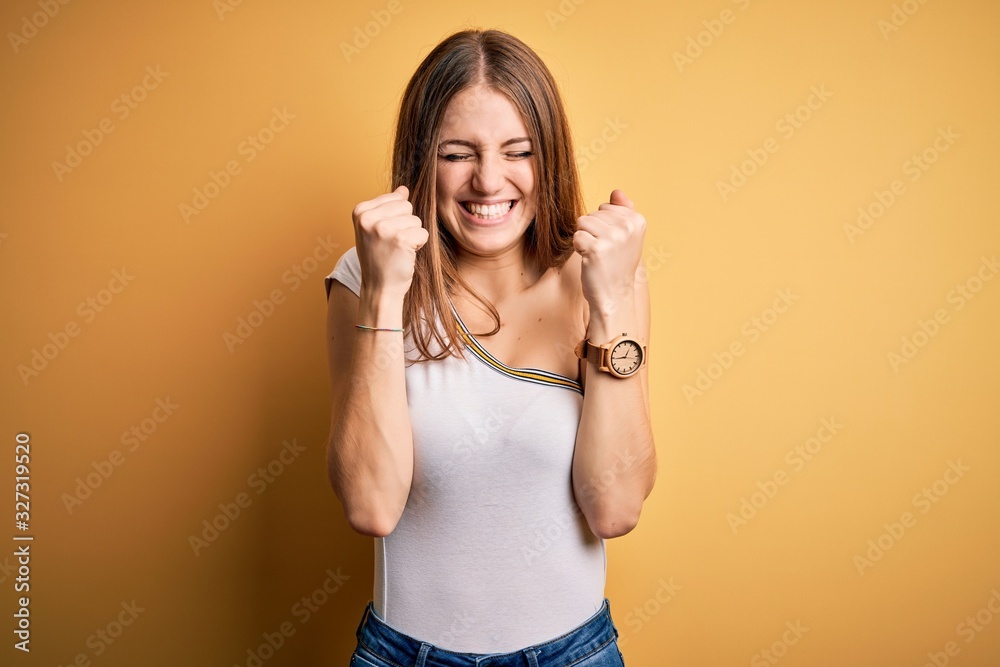 Young beautiful redhead woman wearing casual t-shirt over isolated yellow background excited for success with arms raised and eyes closed celebrating victory smiling. Winner concept. Young beautiful redhead woman wearing casual t-shirt over isolated yellow background excited for success with arms raised and eyes closed celebrating victory smiling. Winner concept.
