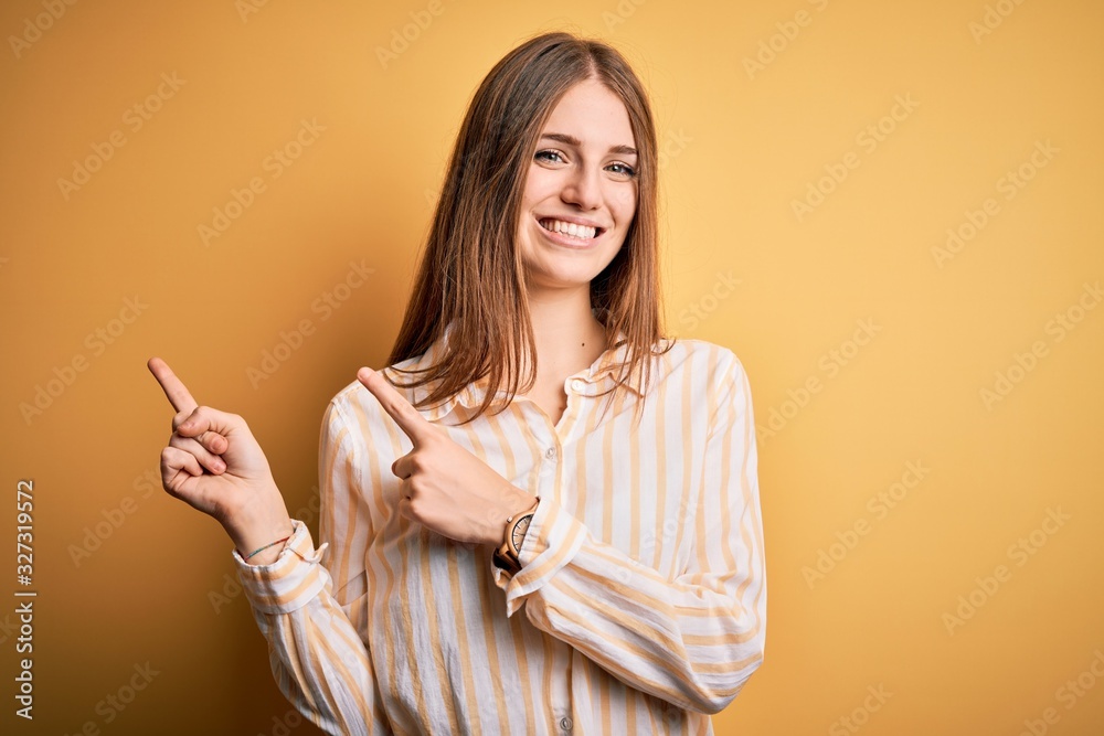 Young beautiful redhead woman wearing casual striped shirt over isolated yellow background smiling and looking at the camera pointing with two hands and fingers to the side. Young beautiful redhead woman wearing casual striped shirt over isolated yellow background smiling and looking at the camera pointing with two hands and fingers to the side.