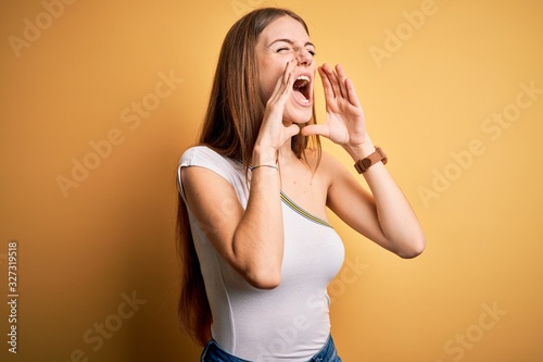 Young beautiful redhead woman wearing casual t-shirt over isolated yellow background Shouting angry out loud with hands over mouth
