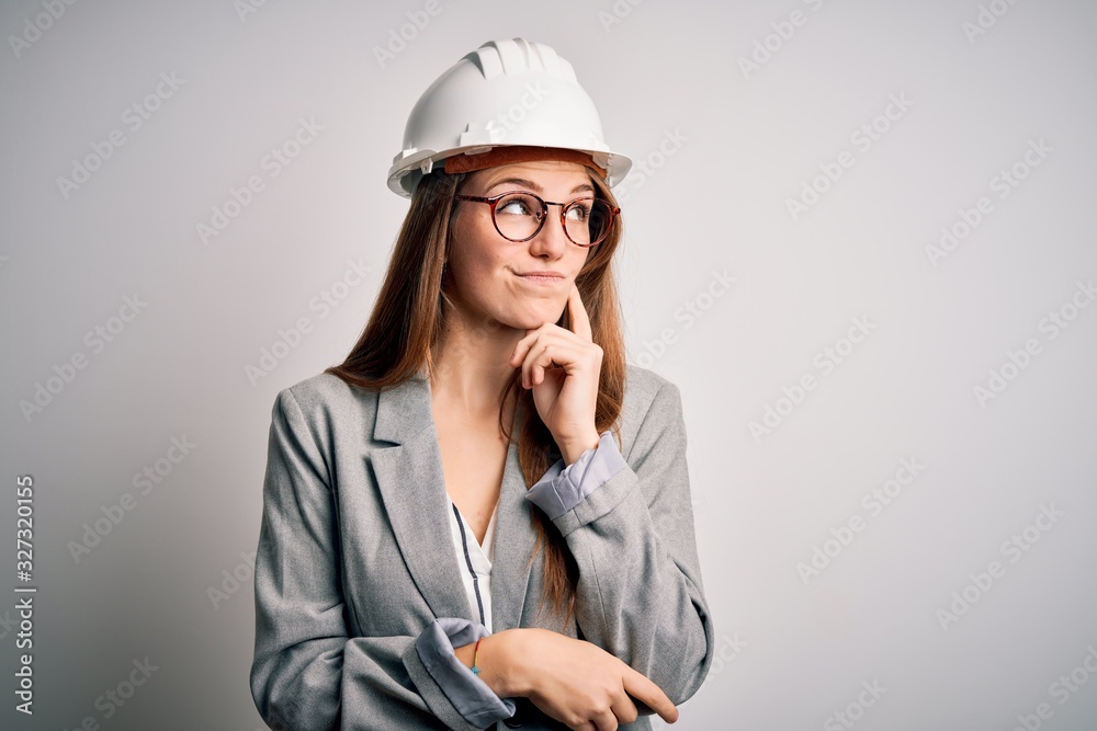 Young beautiful redhead architect woman wearing security helmet over white background with hand on chin thinking about question, pensive expression. Smiling with thoughtful face. Doubt concept. Young beautiful redhead architect woman wearing security helmet over white background with hand on chin thinking about question, pensive expression. Smiling with thoughtful face. Doubt concept.