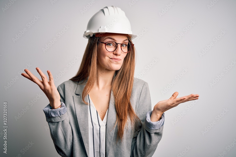 Young beautiful redhead architect woman wearing security helmet over white background clueless and confused expression with arms and hands raised. Doubt concept. Young beautiful redhead architect woman wearing security helmet over white background clueless and confused expression with arms and hands raised. Doubt concept.