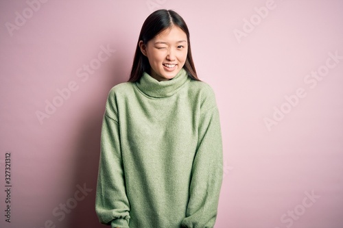 Young beautiful asian woman wearing green winter sweater over pink solated background winking looking at the camera with sexy expression, cheerful and happy face. © Krakenimages.com
