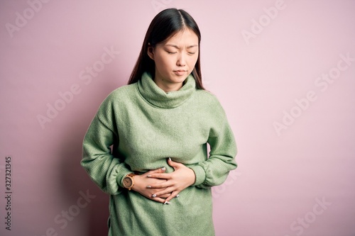 Young beautiful asian woman wearing green winter sweater over pink solated background with hand on stomach because indigestion, painful illness feeling unwell. Ache concept.