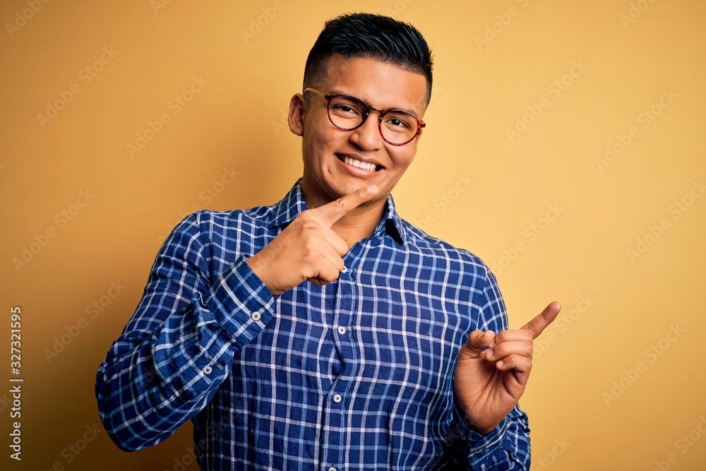 Young handsome latin man wearing casual shirt and glasses over yellow background smiling and looking at the camera pointing with two hands and fingers to the side.