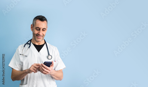 Handsome middle aged male doctor wearing white coat is typing on mobile phone in front of blue wall