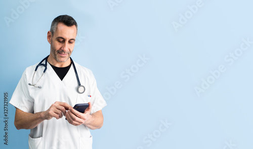 Handsome middle aged male doctor wearing white coat is typing on mobile phone in front of blue wall