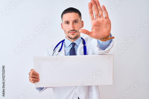 Young doctor man holding blank advertising banner over isolated background with open hand doing stop sign with serious and confident expression, defense gesture