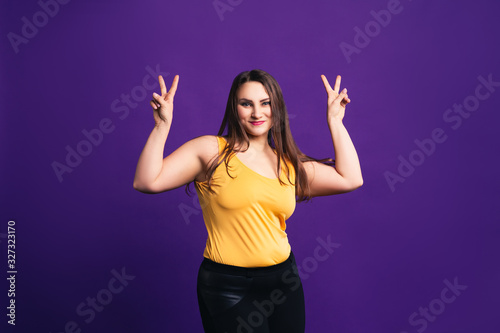 Happy plus size fashion model in casual clothes shows V sign, fat woman on purple background
