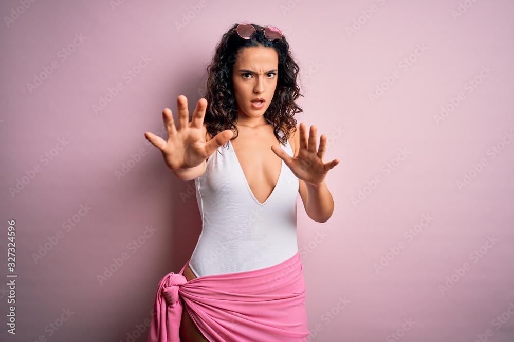Beautiful woman with curly hair on vacation wearing white swimsuit over pink background doing stop gesture with hands palms, angry and frustration expression Beautiful woman with curly hair on vacation wearing white swimsuit over pink background doing stop gesture with hands palms, angry and frustration expression