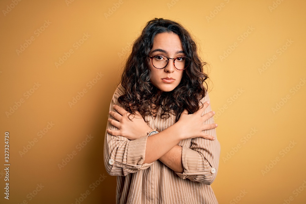 Beautiful woman with curly hair wearing striped shirt and glasses over yellow background shaking and freezing for winter cold with sad and shock expression on face Beautiful woman with curly hair wearing striped shirt and glasses over yellow background shaking and freezing for winter cold with sad and shock expression on face