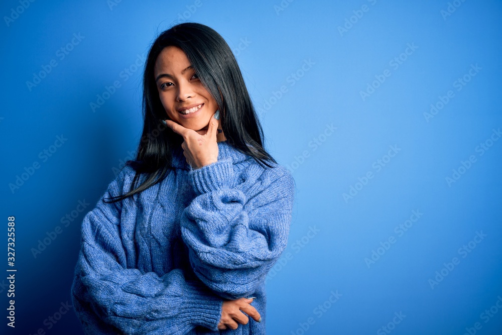 Young beautiful chinese woman wearing casual sweater over isolated blue background looking confident at the camera smiling with crossed arms and hand raised on chin. Thinking positive. Young beautiful chinese woman wearing casual sweater over isolated blue background looking confident at the camera smiling with crossed arms and hand raised on chin. Thinking positive.