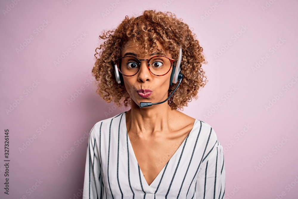 African american curly call center agent woman working using headset over pink background making fish face with lips, crazy and comical gesture. Funny expression.