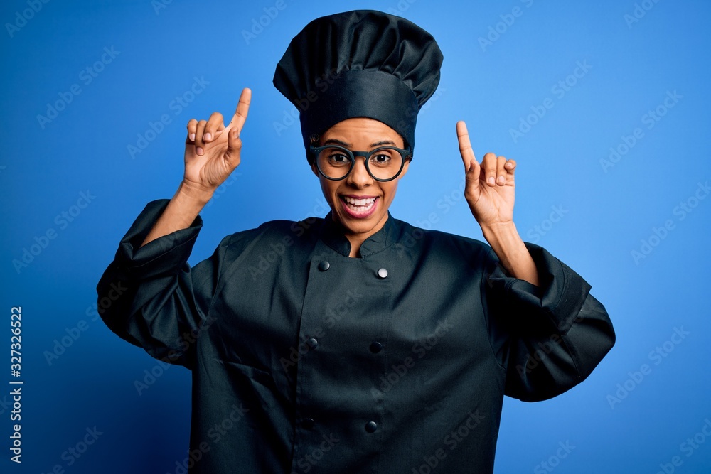 Young african american chef woman wearing cooker uniform and hat over blue background smiling amazed and surprised and pointing up with fingers and raised arms. Young african american chef woman wearing cooker uniform and hat over blue background smiling amazed and surprised and pointing up with fingers and raised arms.