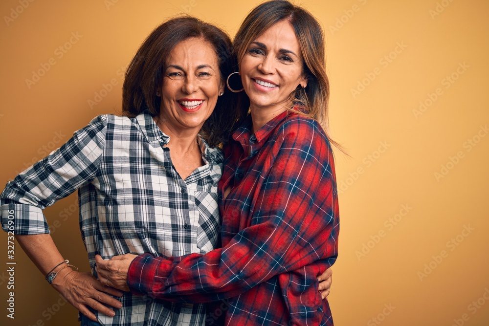 Middle age beautiful couple of sisters smilling happy and confident. Standing with smile on face hugging over isolated yellow background Middle age beautiful couple of sisters smilling happy and confident. Standing with smile on face hugging over isolated yellow background