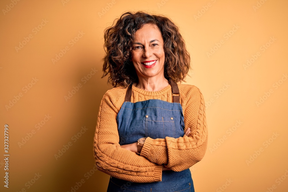Middle age beautiful baker woman wearing apron standing over isolated yellow background happy face smiling with crossed arms looking at the camera. Positive person. Middle age beautiful baker woman wearing apron standing over isolated yellow background happy face smiling with crossed arms looking at the camera. Positive person.