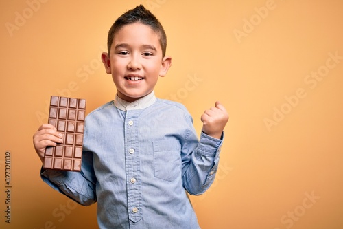 Young little boy kid eating sweet chocolate bar for dessert over isolated yellow background screaming proud and celebrating victory and success very excited, cheering emotion