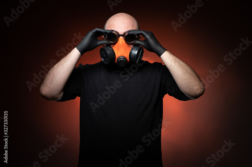 A man in a black t-shirt and rubber gloves puts on a mask and round glasses. A man in a protective mask and round glasses. A man wearing a respirator and glasses on a red-brown background.