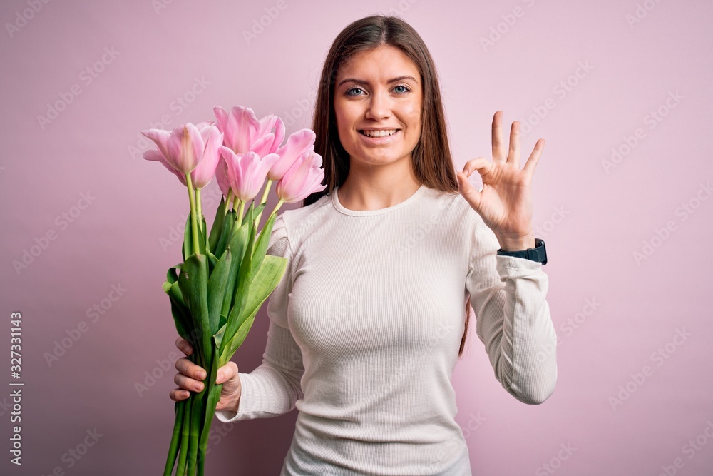 Young beautiful romantic woman with blue eyes holding beauty bouquet of pink tulips doing ok sign with fingers, excellent symbol Young beautiful romantic woman with blue eyes holding beauty bouquet of pink tulips doing ok sign with fingers, excellent symbol