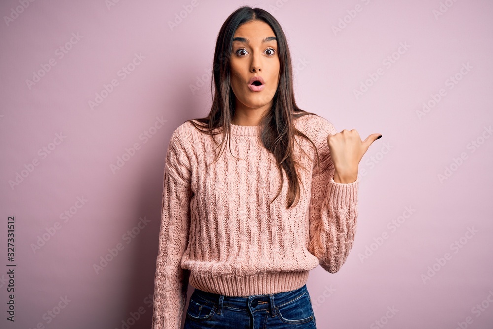 Young beautiful brunette woman wearing casual sweater over isolated pink background Surprised pointing with hand finger to the side, open mouth amazed expression. Young beautiful brunette woman wearing casual sweater over isolated pink background Surprised pointing with hand finger to the side, open mouth amazed expression.