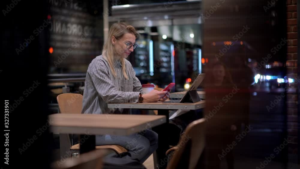 Millennial Caucasian hipster blogger in glasses using smartphone for online networking and chatting with social web followers, evening city lights reflected in coffee shop glass window Millennial Caucasian hipster blogger in glasses using smartphone for online networking and chatting with social web followers, evening city lights reflected in coffee shop glass window