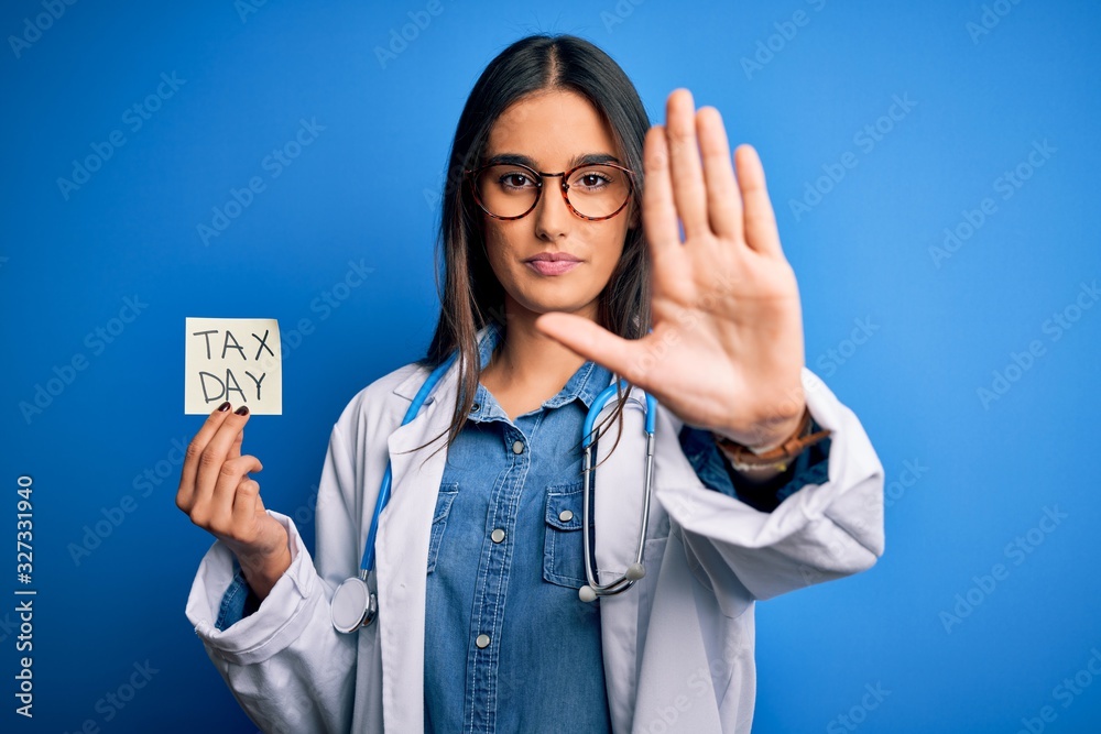 Young doctor woman wearing stethoscope holding paper with tax day message with open hand doing stop sign with serious and confident expression, defense gesture Young doctor woman wearing stethoscope holding paper with tax day message with open hand doing stop sign with serious and confident expression, defense gesture