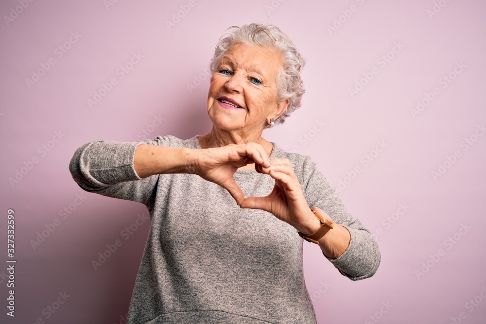 Senior beautiful woman wearing casual t-shirt standing over isolated pink background smiling in love doing heart symbol shape with hands. Romantic concept. - obrazy, fototapety, plakaty 