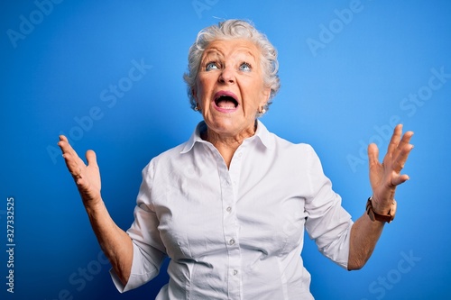 Senior beautiful woman wearing elegant shirt standing over isolated blue background crazy and mad shouting and yelling with aggressive expression and arms raised. Frustration concept.