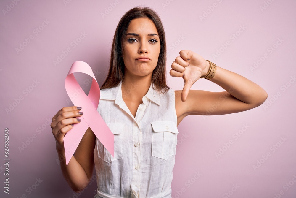 Young beautiful brunette woman holding cancer ribbon symbol over pink background with angry face, negative sign showing dislike with thumbs down, rejection concept Young beautiful brunette woman holding cancer ribbon symbol over pink background with angry face, negative sign showing dislike with thumbs down, rejection concept