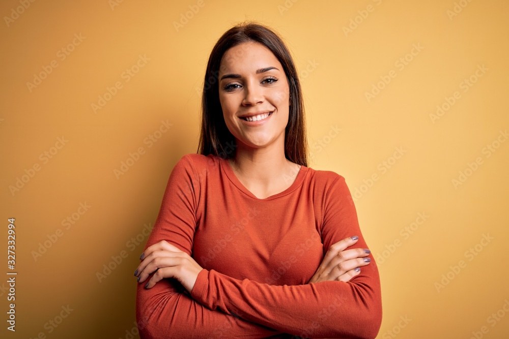 Obraz premium Young beautiful brunette woman wearing casual t-shirt standing over yellow background happy face smiling with crossed arms looking at the camera. Positive person.
