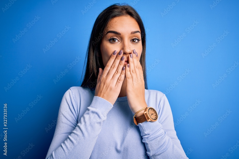 Young beautiful brunette woman wearing casual sweater standing over blue background laughing and embarrassed giggle covering mouth with hands, gossip and scandal concept